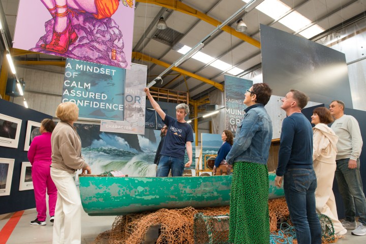 Group of people observing art displays in an industrial-style exhibition space.