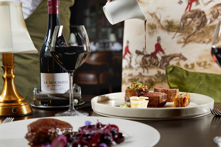 Table with wine, steak served on a plate, and sauce being poured from a jug in a restaurant setting.