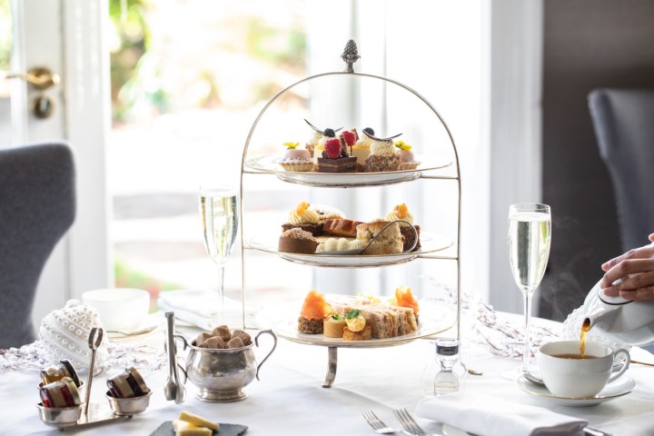 Three-tiered afternoon tea stand with pastries, sandwiches, and champagne on a table.