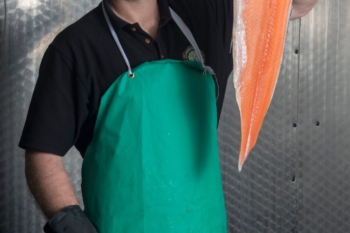 Man in green apron holding a large salmon fillet in a fish market.