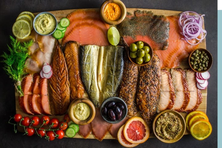 A deli board with various smoked fish, lemon slices, and condiments on a dark surface.
