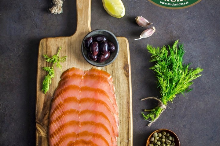 Sliced salmon on a wooden board with olives, capers, lemon, garlic, dill, and bread.