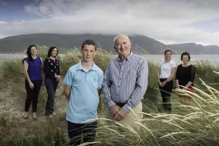 Group of six people standing in grassy dunes with mountains and sea in background.