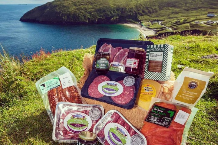 A variety of packaged Irish foods are displayed on grass by a coastal landscape with hills and sea.
