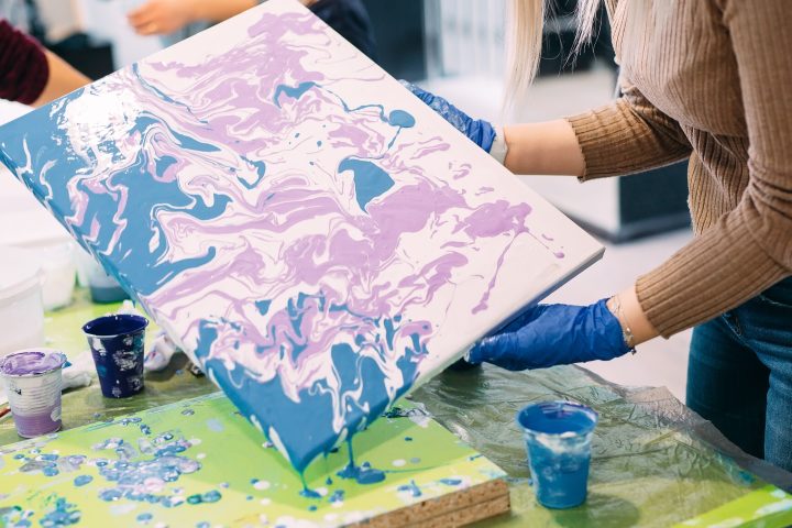 Person tilting canvas with blue and lavender paint, art supplies on table.