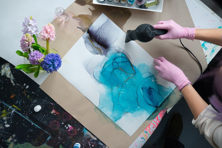 Person using heat gun on blue abstract painting, nearby are flowers and paints.
