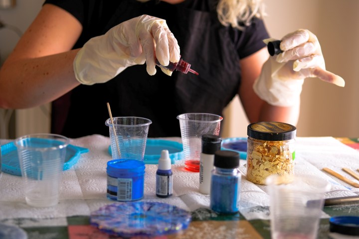 Person wearing gloves mixes colorful materials at a table with art supplies.