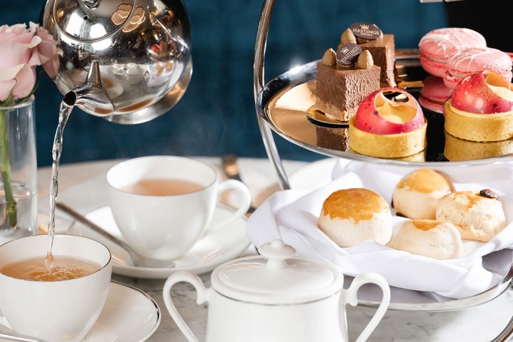 Afternoon tea setting with pouring tea, tiered dessert stand, and pastries on table.