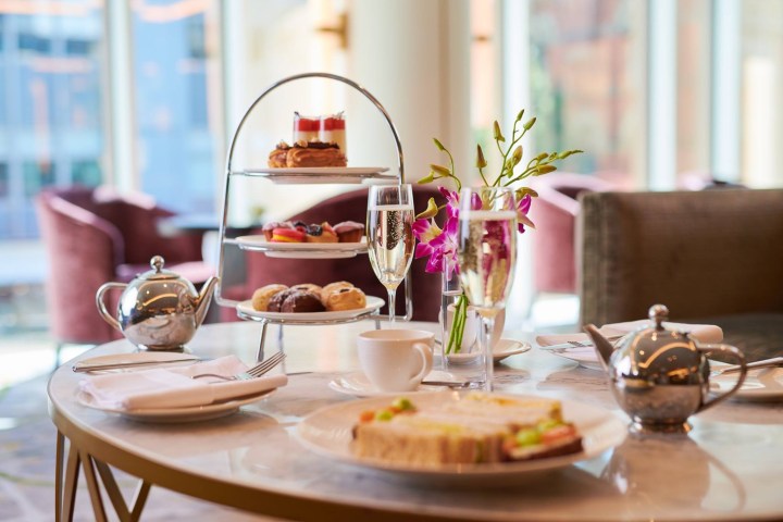 Elegant afternoon tea setup with tiered tray, teapot, champagne, and flowers on a marble table in a bright room.