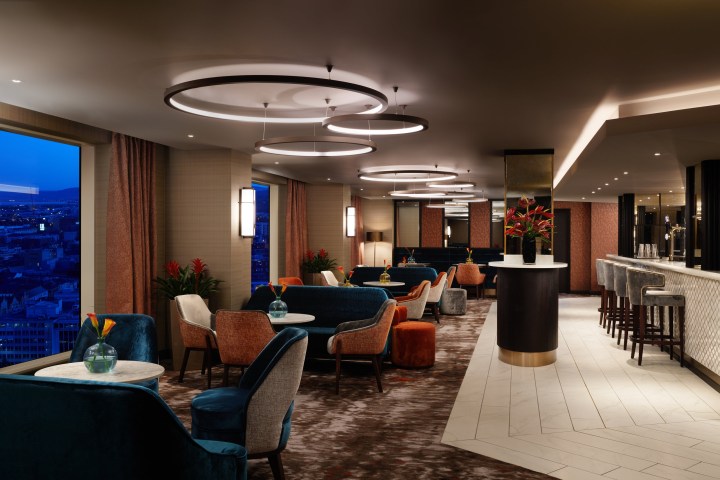 Modern lounge with elegant seating, circular lights, city view, and a bar with stools.