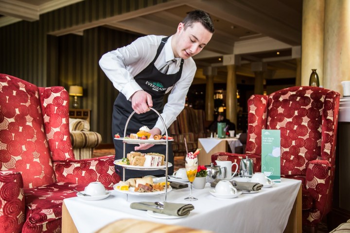 Waiter serves afternoon tea with tiered tray of sandwiches and pastries in a cozy room.