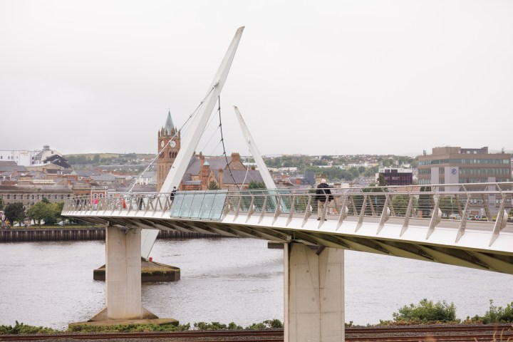 Modern pedestrian bridge over river with cityscape in background under cloudy sky.