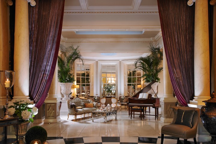 Elegant living room with grand piano, luxurious seating, large plants, and tall, maroon drapes.