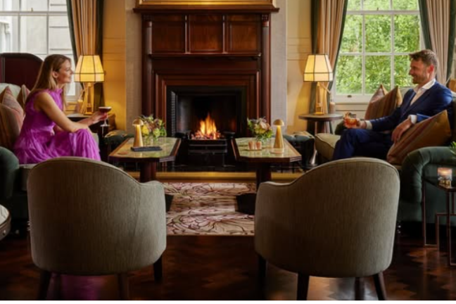 Two people talking in a cozy living room with a lit fireplace and elegant decor.