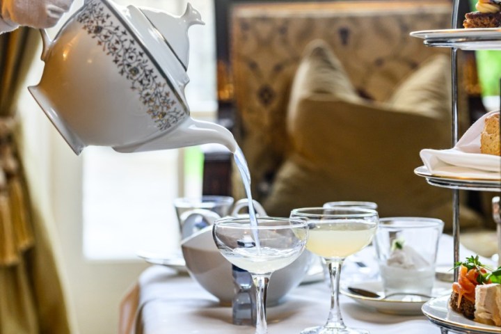 Afternoon tea setting with teapot pouring, sweets, and sandwiches on a table.