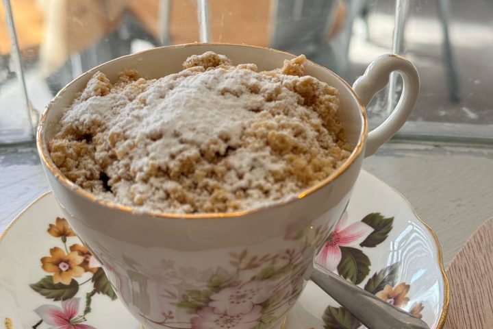 Cup with crumble dessert on floral saucer and spoon, by window.