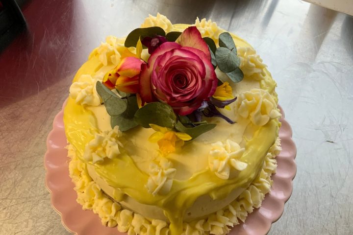 Cake with yellow icing, pink rose, and flowers on a pink plate on a metal surface.