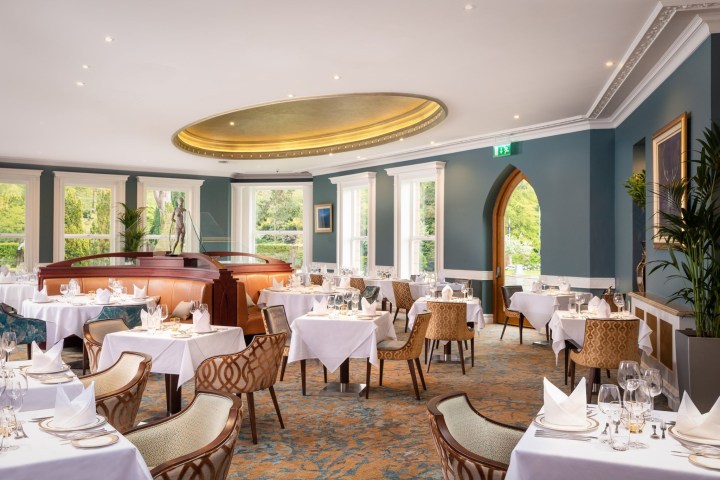Elegant restaurant with round tables, white tablecloths, and teal walls, featuring a large oval ceiling design.