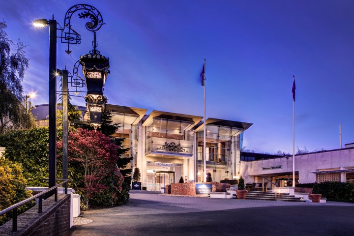 Elegant hotel entrance at dusk with lit signage and flags, surrounded by greenery.