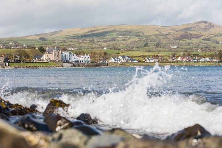 Waves crashing on rocks with a coastal village and hills in the background.