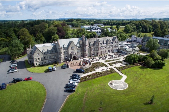 Aerial view of a large stone hotel with parked cars and green lawns surrounding it.