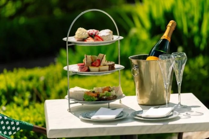 Afternoon tea setup with tiered tray, champagne in ice bucket, and flutes on an outdoor table.