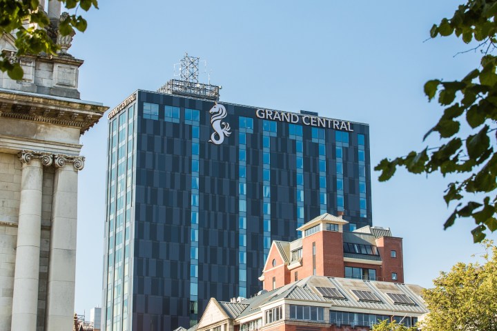 Tall building labeled Grand Central beside older architecture and trees, under blue sky.