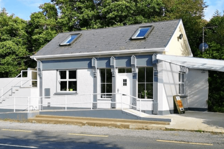 Small white building with grey roof, large windows, and trees in background.
