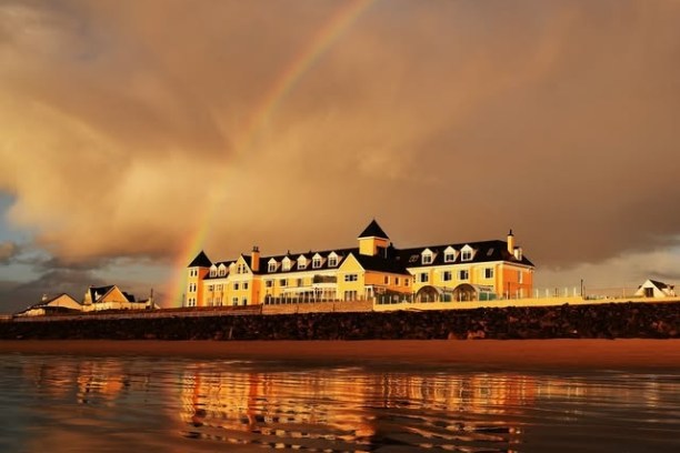 Large building by the beach with a rainbow in the cloudy sky, reflections on wet sand.