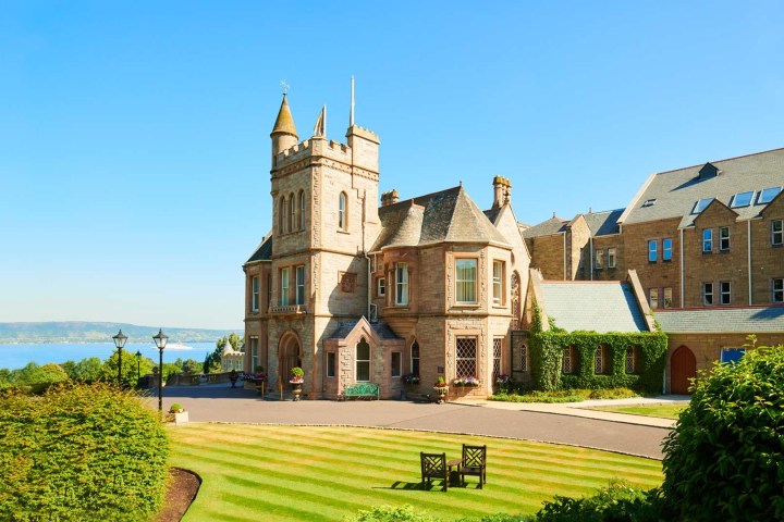 Castle-like building with turrets, manicured lawns, and a view of water and hills in the background.