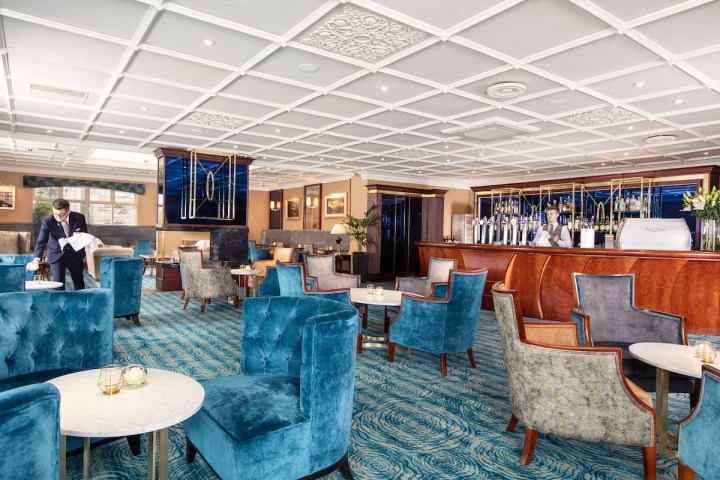 Elegant hotel lounge with blue velvet chairs, marble tables, and a bar counter being served by staff.