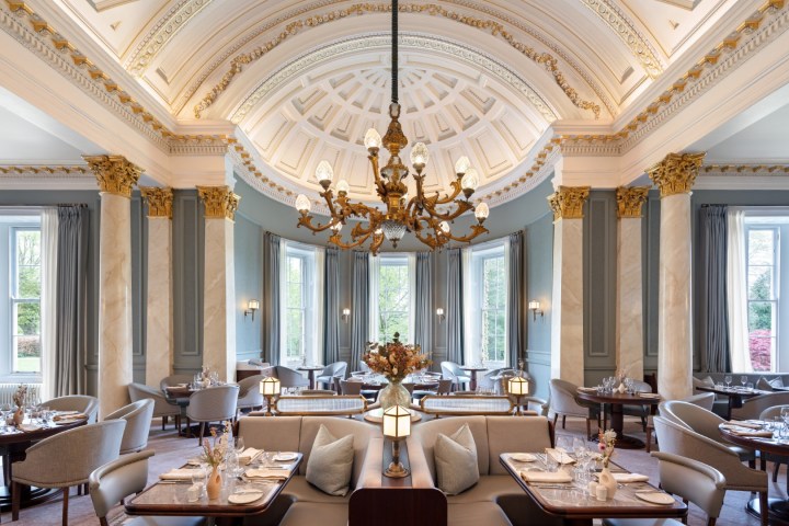 Elegant restaurant with high ornate ceiling, chandelier, columns, large windows, and set tables.