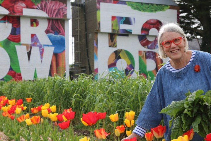 Smiling woman in garden, holding greens, with colorful mural saying 'GROW' in the background.