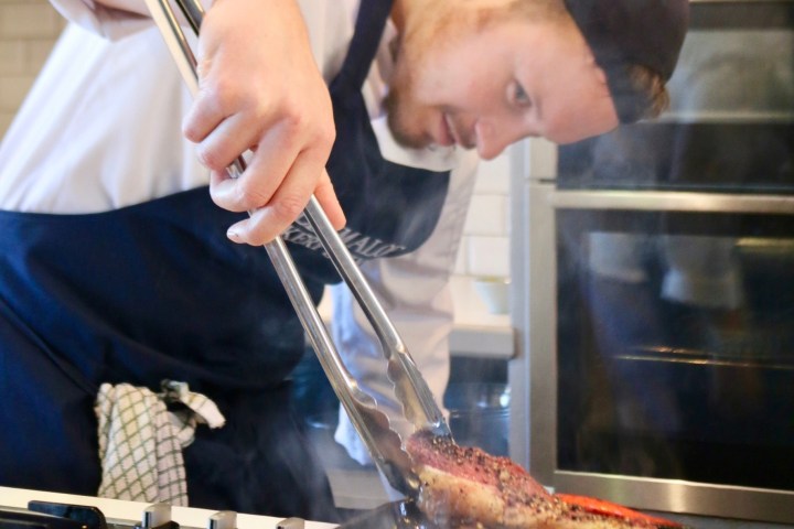 Chef cooking steaks on a grill pan with tongs, wearing a black cap and apron.