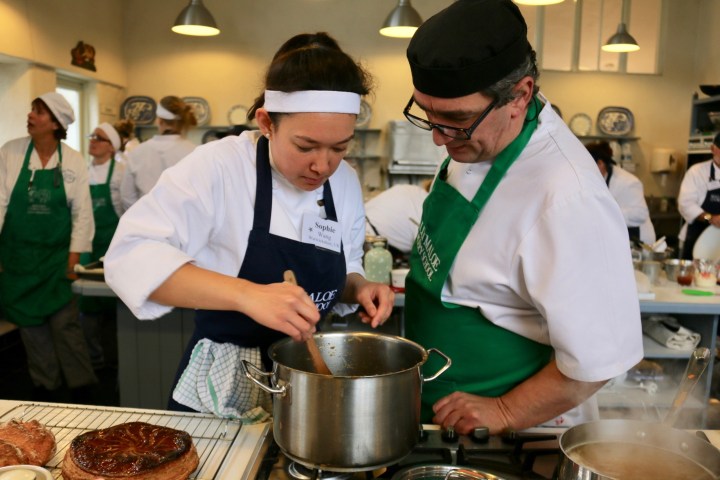 Two chefs in a kitchen, with one stirring a pot on a stove.