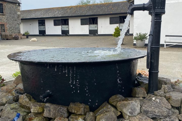Old-fashioned water pump filling a large basin with stone surroundings and buildings in the background.