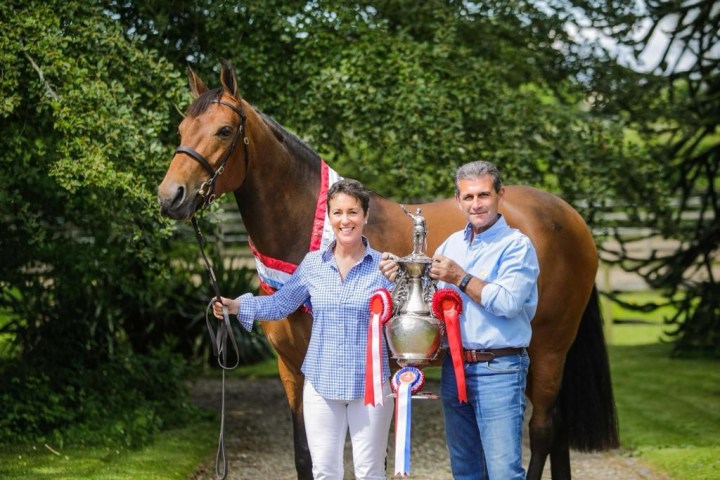 Two people stand with a horse, holding a trophy with red ribbons, outside in a grassy area.