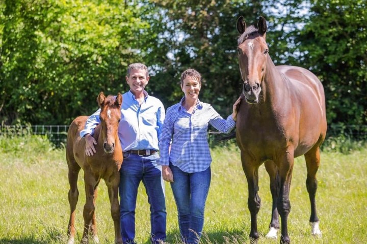 Two people smiling with a foal and a horse in a sunny field.