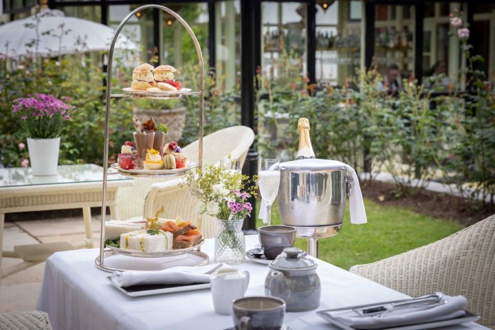 Outdoor table set with tiered afternoon tea, champagne, and flowers in garden setting.