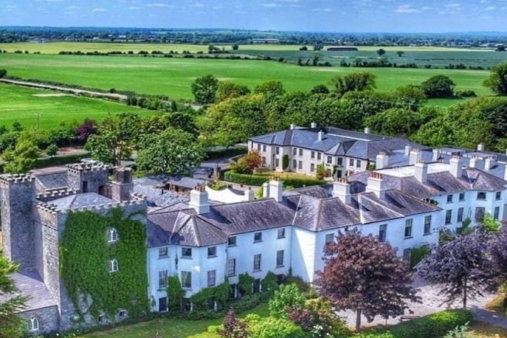 Aerial view of a historic manor with ivy-covered walls, surrounded by lush greenery and fields.