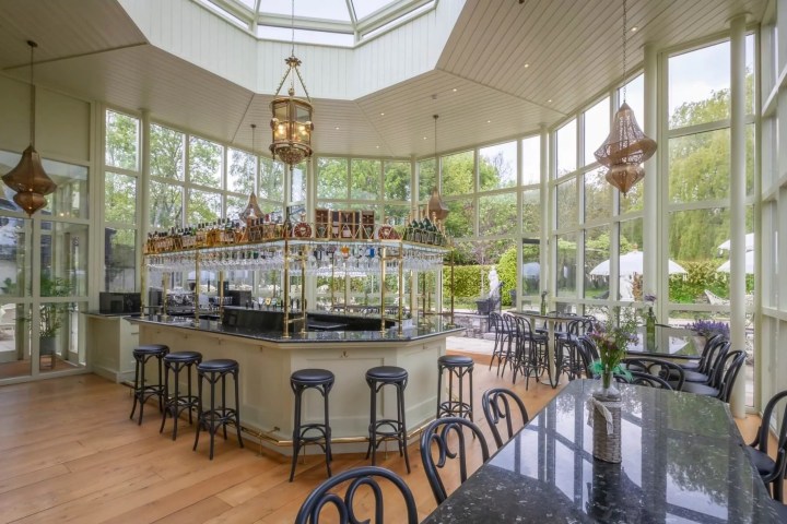 Spacious glass-walled bar with central counter, stools, and large windows showcasing greenery outside.