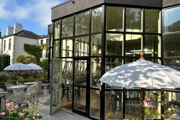 Glass-walled building with outdoor seating, umbrellas, and pink flowers in a garden setting.