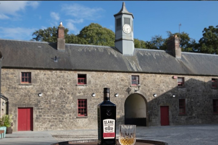 Stone building with clock tower, bottle of Slane whiskey and glass on barrel in foreground.