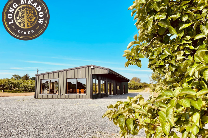 Metal building with large windows, gravel lot, green trees, and blue sky, labeled 'LONG MEADOW CIDER' in corner.