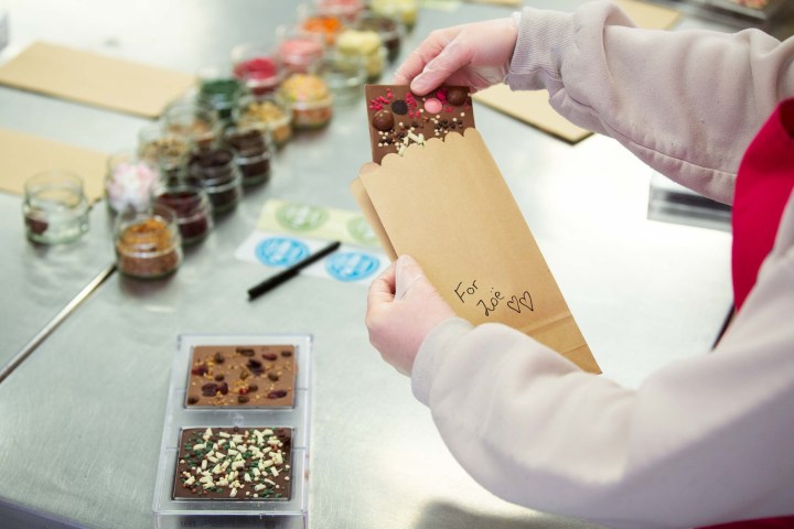 Person placing decorated chocolate bar into a kraft paper bag labeled 'For Zoe' on a table with ingredients.