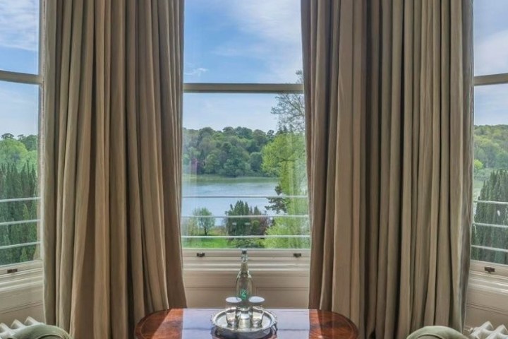 Table by tall windows with lake and trees view, flanked by beige curtains.