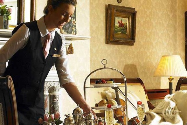 Waitress serving tea in an elegant room with ornate decor and pastries on a tiered tray.