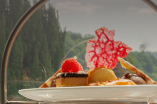 Plate of desserts with red garnish in arched stand, green trees in background.