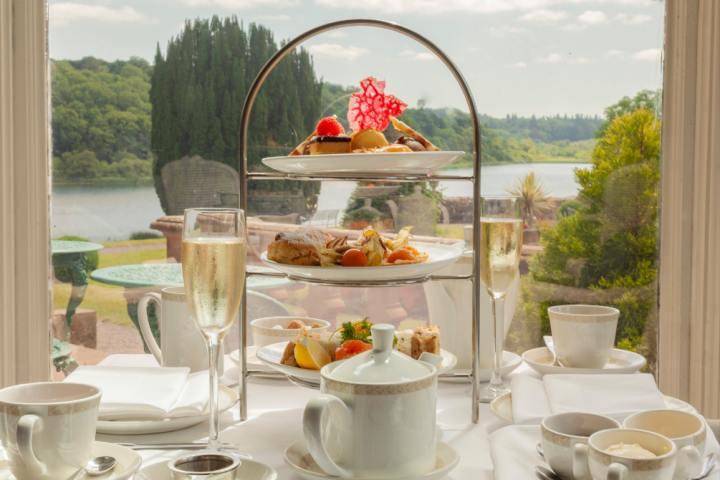 Afternoon tea setup with tiered stand, tea, and champagne by a window overlooking a scenic view.