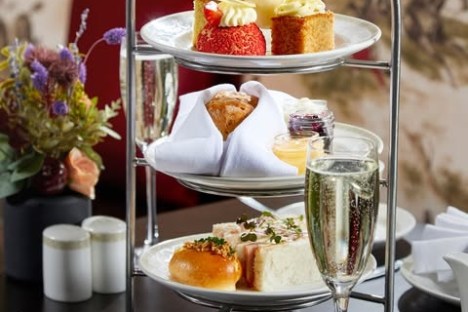 Three-tiered stand with assorted pastries, sandwiches, tea, and a glass of champagne on a table.
